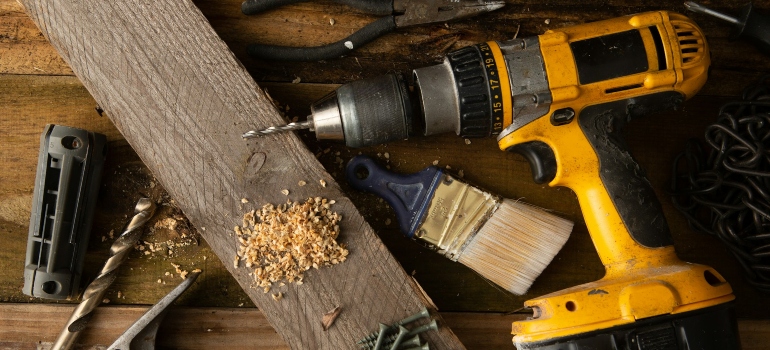 A variety of tools, including a hammer and cordless drill, sit on top of a table made of wood boards.