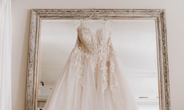 Stylish white bridal dress hanging on wall with mirror before wedding celebration