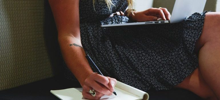 A woman typing on a laptop and writing in a notebook simultaneously.