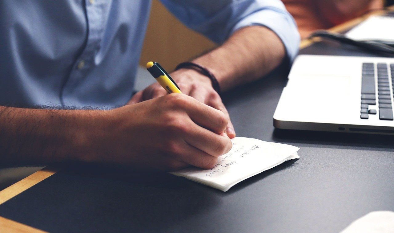A person writing something in a notepad next to a laptop, representing the process of coordinating large-scale moves.
