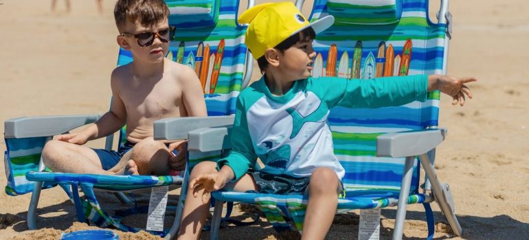 Two boys are sitting in chairs on a sandy beach.