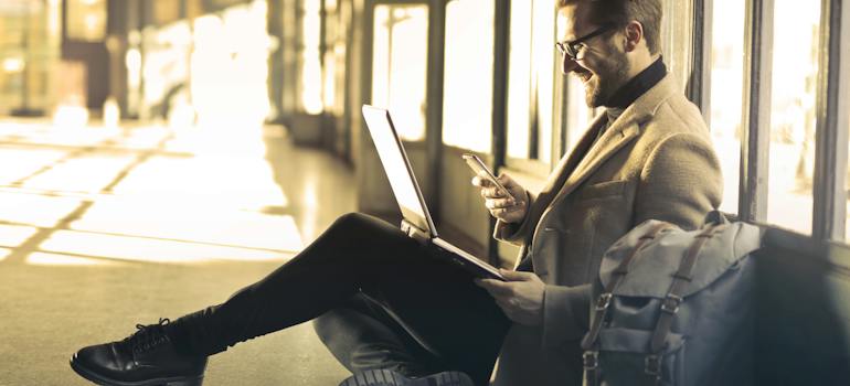 A man texting on a smartphone and holding a laptop