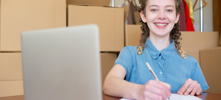 A Woman in a Blue Denim Shirt sitting among boxes and considering portable storage or traditional units