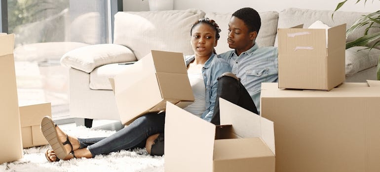 A young couple sitting in a living room full of cardboard boxes