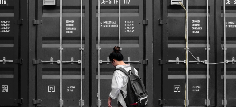 Photography of Woman in Gray Long Sleeve Shirt Carrying Black Backpack Standing in Front of Black Intermodal Container and thinking about the perks of portable storage containers